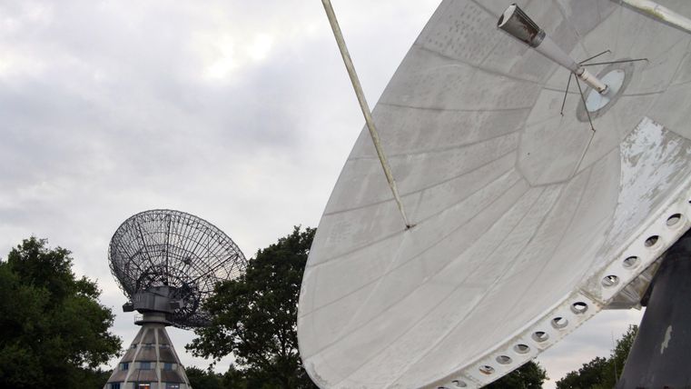 Zwei Satellitenschüsseln stehen in einem offenen Gelände. Der Himmel ist bewölkt und die Umgebung besteht aus Bäumen.