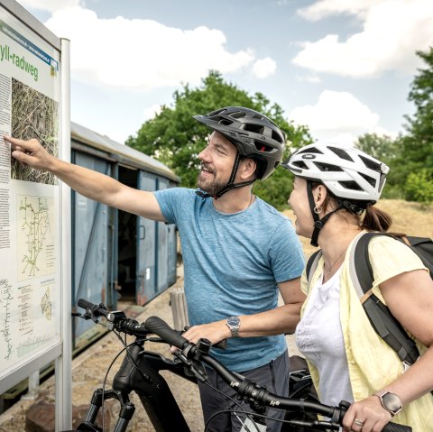 Deux cyclistes casqu&eacute;s &eacute;tudient un panneau d'information sur la piste cyclable de la Kyll. Ils sourient et montrent la carte. En arri&egrave;re-plan, des arbres et un conteneur bleu., &copy; Eifel Tourismus GmbH, Dominik Ketz