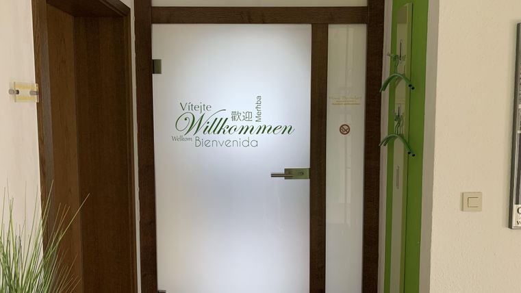 Eine Eingangstür mit der Aufschrift "Willkommen" in verschiedenen Sprachen. Die Tür hat einen modernen Rahmen aus Holz und transparente Glaselemente.