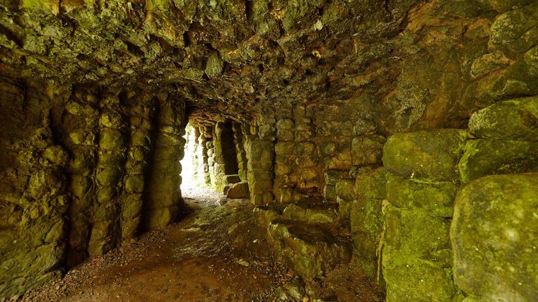 Een smalle tunnel omgeven door rotsen met met mos bedekte muren. Aan het einde van de tunnel is fel licht zichtbaar.