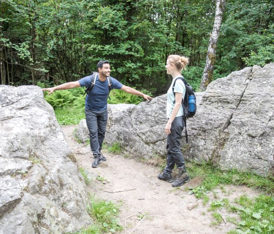 Twee wandelaars staan tussen grote rotsen in het bos en kletsen. Ze dragen rugzakken en wandelkleding., © Eifel-Tourismus GmbH, shapefruit AG A. Röser