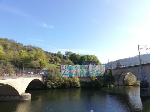 Eine Brücke über einen ruhigen Fluss mit bunten Wandmalereien im Hintergrund. Grüne Bäume und ein klarer blauer Himmel runden die Szene ab.