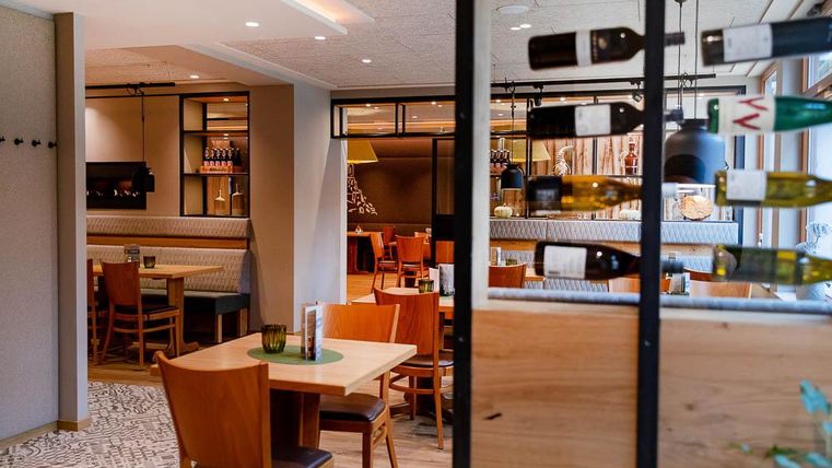 Ein einladendes Restaurant mit Holzstühlen und Tischen. Im Hintergrund sind Weinflaschen und eine gemütliche Atmosphäre zu sehen.
