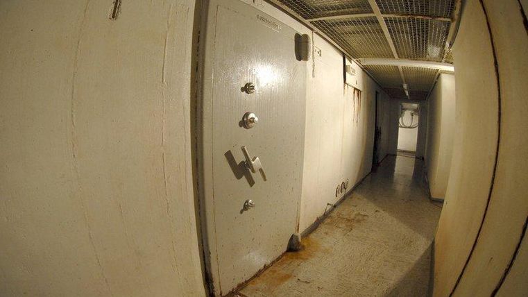 A narrow corridor with a solid, locked door at the end. The floor appears dirty and the walls are plain and painted white.