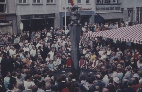 Eine große Menschenmenge versammelt sich um einen Brunnen mit einer Statue in der Mitte. Im Hintergrund sind Markisen und Geschäfte zu sehen.