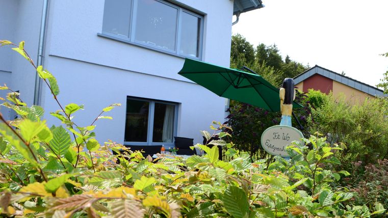 A modern house with a green sunshade and a garden sign. In front, various plants and shrubs are growing.