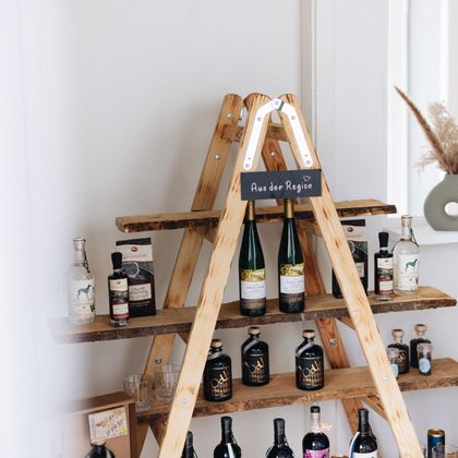 Ein Holzregal mit verschiedenen Flaschen steht in einem modernen Raum. Im Hintergrund sind dekorative Elemente wie ein graues Keramikgefäß und getrocknete Pflanzen zu sehen.