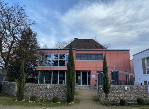 Ein modernes Gebäude mit einer pinken Fassade und großen Fenstern. Der Eingangsbereich ist von Bäumen und einem Steinzaun umgeben.