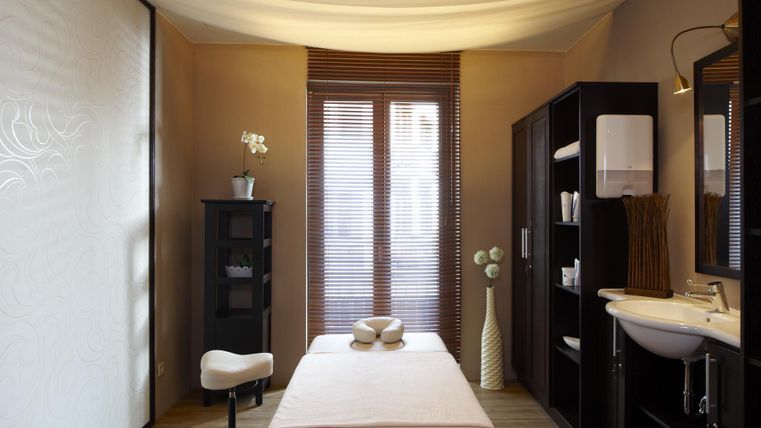 Een rustige wellnessruimte met een massagetafel en zachte kleuren. Er zijn ramen met jaloezieën en een elegante decoratie.