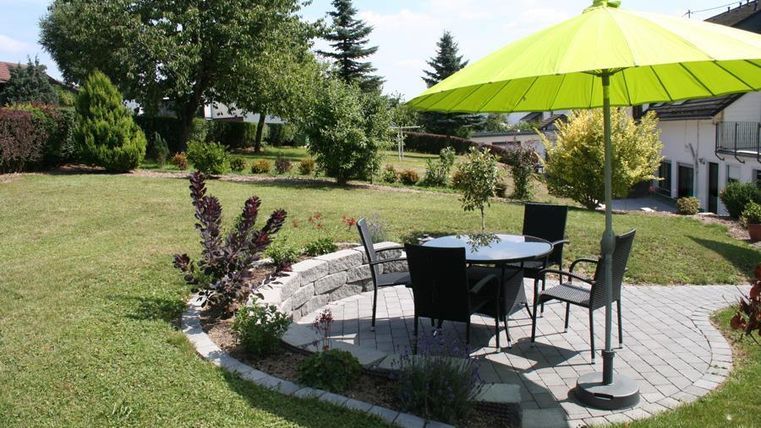 An inviting garden with a round stone terrace, a table, and chairs. A large, brightly colored green parasol provides shade.