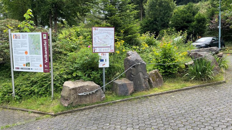 Een entree met informatieborden en stenen, omringd door groene struiken en bomen. Op de achtergrond is een parkeerplaats te zien.
