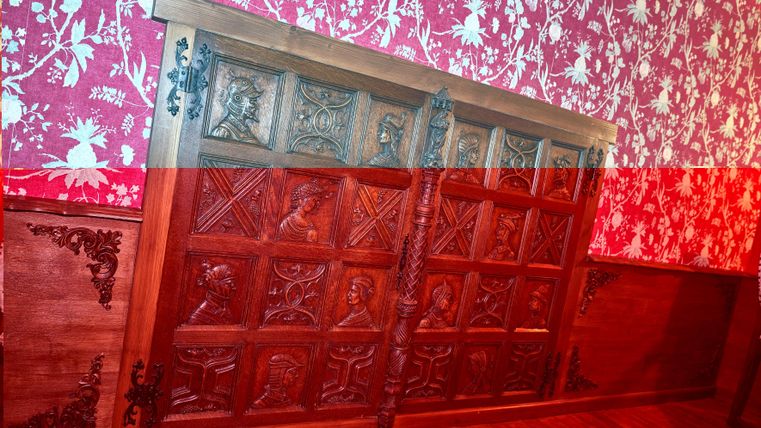An intricately carved wooden panel with various reliefs. It is mounted on a wall with red-and-white floral wallpaper.