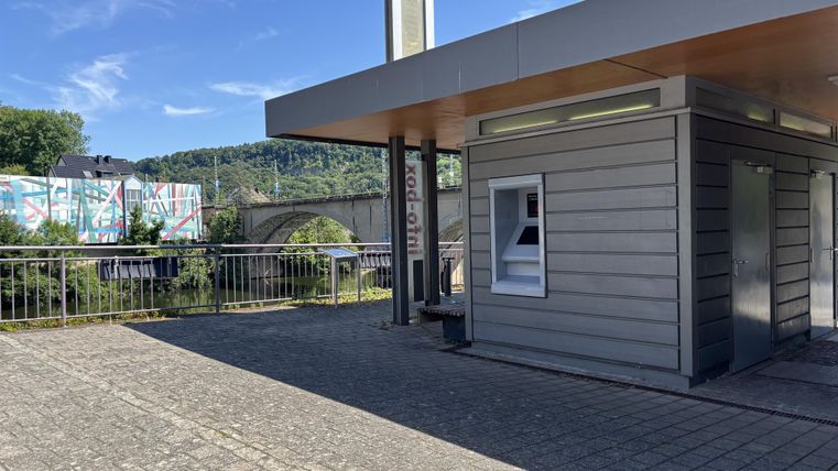 Een moderne kiosk van hout en glas staat op een geplaveid plein. Op de achtergrond is een brug en kleurrijke graffiti te zien.