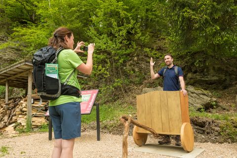 Eine Frau fotografiert einen Mann, der hinter einem hölzernen Wagen steht, auf einem Wanderweg im Wald.