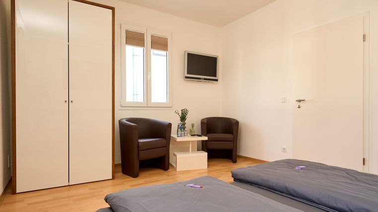 Ein einfaches, modernes Zimmer mit zwei Betten und einem Flachbildfernseher. Es gibt zwei Stühle und einen kleinen Tisch neben einem Schrank.