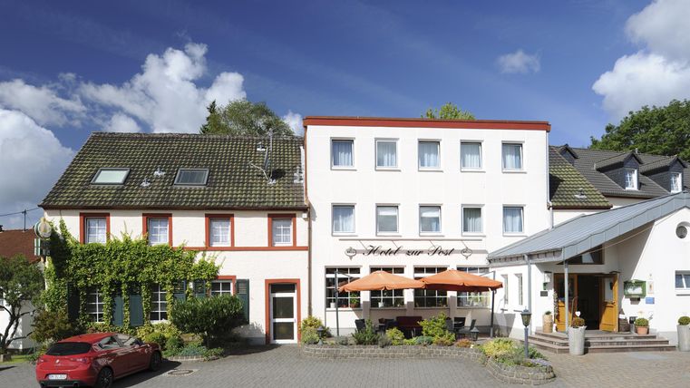 Ein charmantes Hotel mit einem modernen Anbau und gemütlicher Terrasse. Der Himmel ist blau mit einigen Wolken, und es gibt gepflegte Pflanzen an der Fassade.