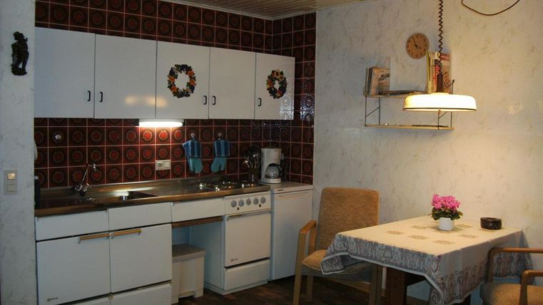 Eine kleine Küche mit weißen Schränken und roten Fliesen. Ein Tisch mit Stühlen und einer Vase mit Blumen steht in der Ecke.