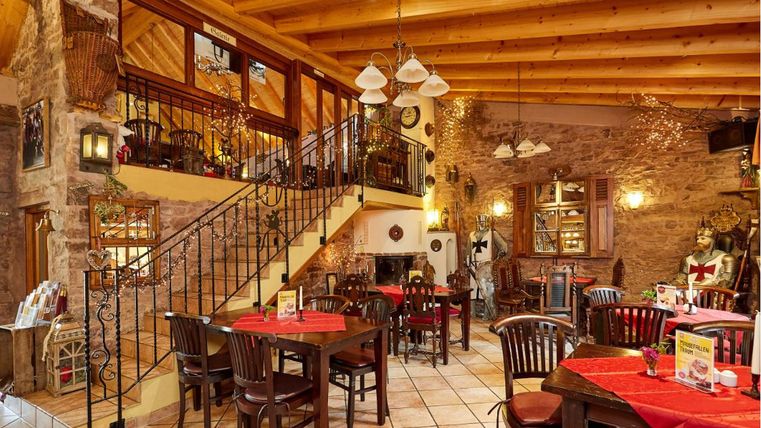 Interior view of a rustic restaurant with wooden beam ceiling, knight armors, and red tablecloths.