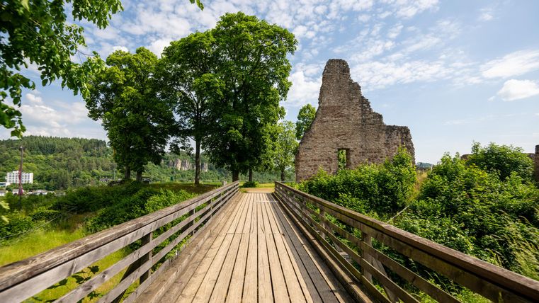 Eine Holzbrücke führt zu einer alten Burgruine, umgeben von üppigem Grün und Bäumen unter einem blauen Himmel mit Wolken.