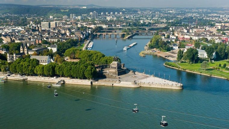 An aerial view of a river with a city in the background. In the foreground, there is a statue and cable cars, surrounded by trees.