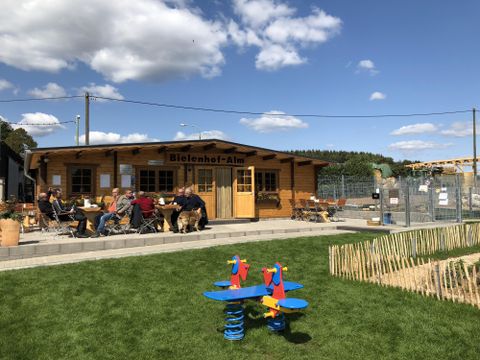Op de achtergrond zie je een grote, eencellige houten hut. In het midden is een dubbele deur. Boven de deur hangt een houten bord met de tekst "Bielenhof Alm". Rechts en links van de deur zijn er vensters met luiken en bloembakken. Voor de hut is er een betegeld terras met enkele tafels en stoelen. Twee van de tafels zijn bezet door een groep mannen. Op de voorgrond staat op een grasveld een wipkip in de kleuren blauw, geel en rood.