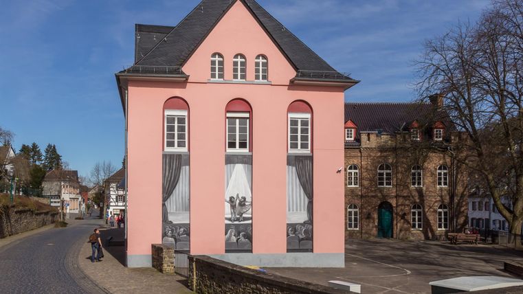 Ein rosa Gebäude mit Bildern von Vorhängen und Menschen an der Fassade. Im Hintergrund sind weitere historische Gebäude und Bäume zu sehen.