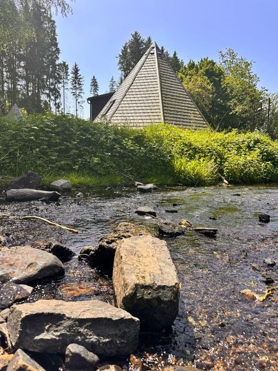 Ein modernes, pyramidenförmiges Gebäude steht in der Natur. Im Vordergrund gibt es einen kleinen Bach und Steine.