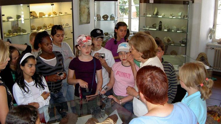 Kindergruppe mit einer älteren Frau im Naturkundemuseum, umgeben von Vitrinen mit Steinen. Die Kinder hören aufmerksam zu.