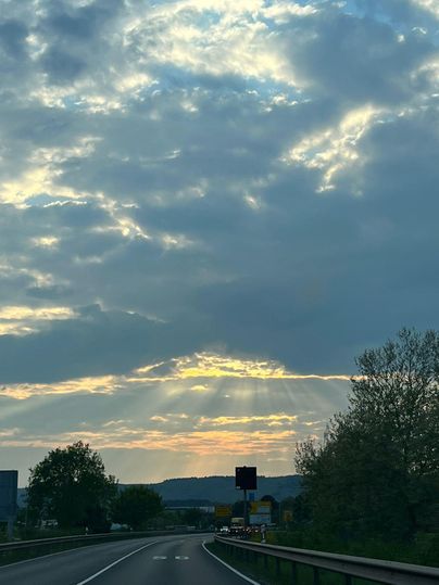 A road that leads through a rural area, under a cloudy sky. Sun rays gently break through the clouds just before sunset.