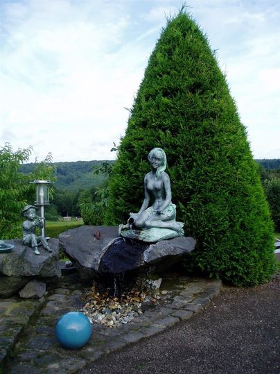 Eine Statue einer sitzenden Figur, die Wasser aus einer Schüssel gießt, steht auf einem großen Stein. Im Hintergrund befindet sich ein hoher, grüner Baum und eine kleine Figur neben der Statue.