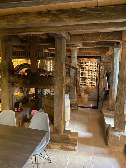 Ein rustikales Weingut mit Holzstrukturen und einem Weinkeller. Im Vordergrund sind ein Tisch und moderne Stühle sichtbar.