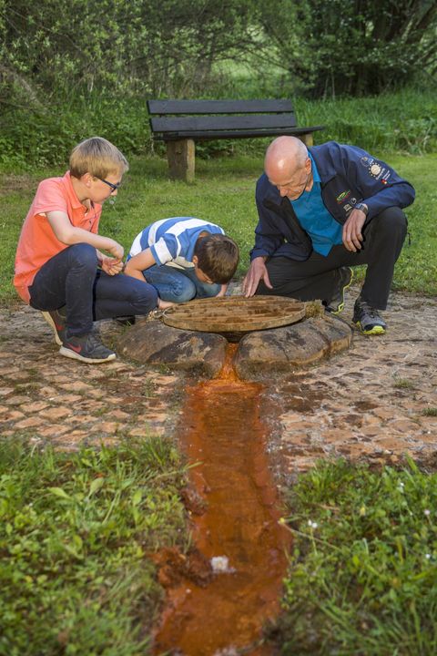 Drei Personen, darunter zwei Kinder und ein Erwachsener, betrachten eine Quelle mit rostfarbenem Wasser im Freien. Im Hintergrund steht eine Bank.