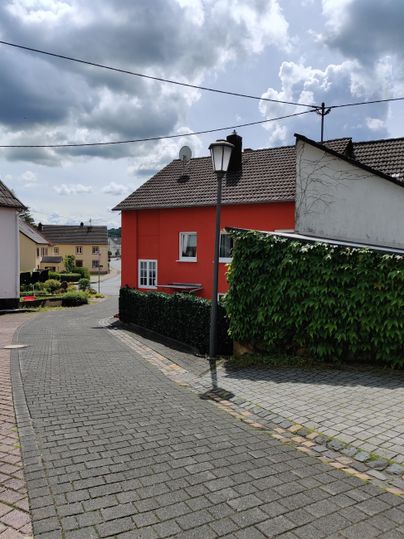 A charming street with a red house and cobblestone pavement. In the background, clouds and some houses can be seen.