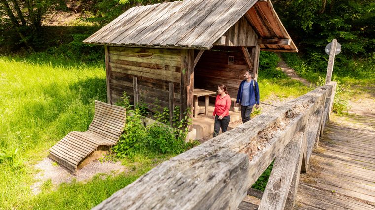 Eine Holzschutzhütte inmitten von grünem Gras und Bäumen. Zwei Personen stehen in der Nähe der Hütte und erkunden die Umgebung.