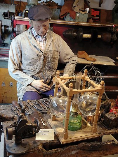 A shoemaker's table with tools and a mannequin in work clothes in a museum. Machines are visible in the background.