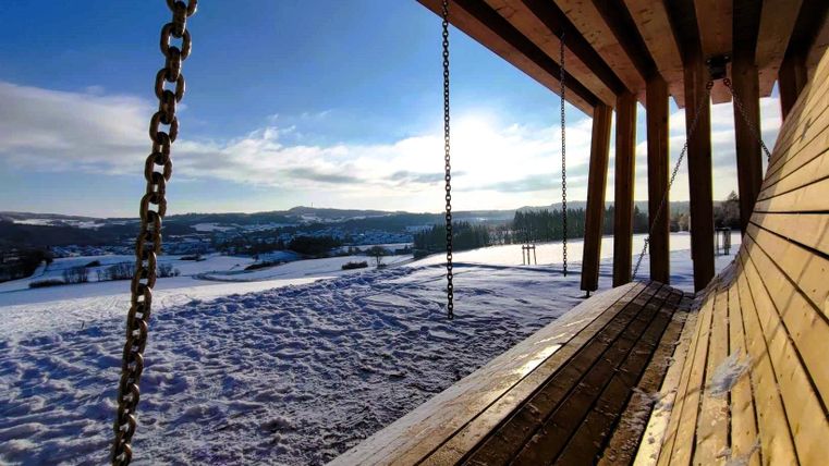 Eine Aussichtsbank aus Holz in einer verschneiten Landschaft. Im Hintergrund sind sanfte Hügel und ein klarer blauer Himmel zu sehen.
