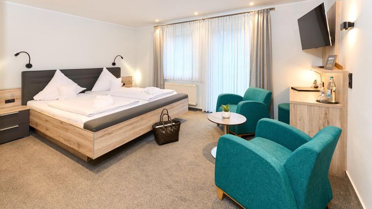 Een moderne hotelkamer met een tweepersoonsbed, een gezellige zithoek en een televisie. Grote ramen zorgen voor veel daglicht en een aangename sfeer.