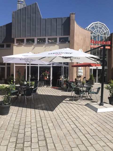 Außenbereich des Bistros mit Tischen, Stühlen und Sonnenschirmen auf einem gepflasterten Platz. Im Hintergrund ein Gebäude mit Glasdach.