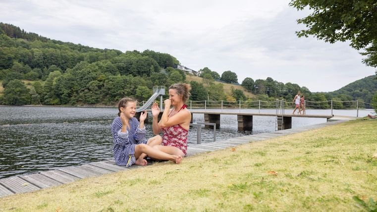 Zwei Mädchen spielen und lachen am Ufer eines Gewässers. Im Hintergrund ist eine Holzbrücke und grüne Hügel zu sehen.