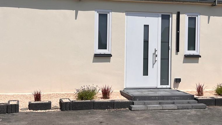 A modern house with a white front door and large windows. In front of the house are well-maintained plants and a beautiful path.