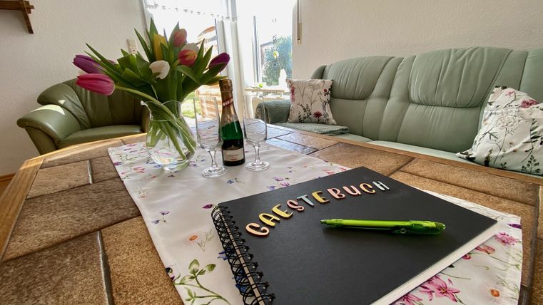 Ein gemütlicher Wohnbereich mit einem Tisch, auf dem ein Gästebuch, ein grüner Stift und eine Flasche Sekt stehen. Im Hintergrund sind frische Tulpen in einer Vase und ein bequemer Sessel zu sehen.