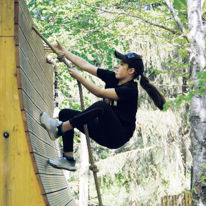 Een meisje klimt op een houten helling in een bosgebied. Ze draagt sportkleding en concentreert zich op de klim.