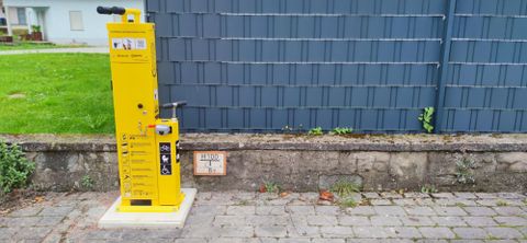 Eine gelbe Fahrradpumpe steht an einer Wand. Der Boden besteht aus Pflastersteinen und es ist grünes Gras im Hintergrund sichtbar.