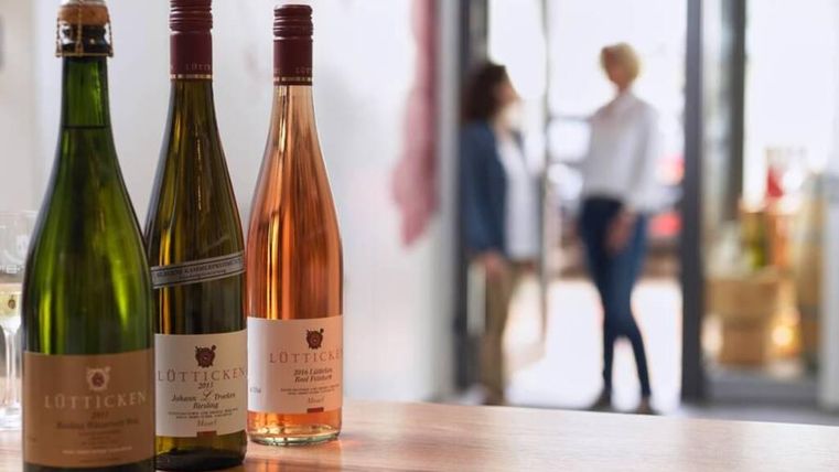 Three bottles of wine are on a wooden table. In the background, there are two people talking to each other.