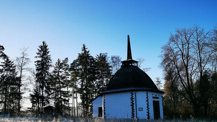 Een klein, wit gebouw met een spitse dak staat in een bevroren veld omringd door bomen. De lucht is helder en blauw.