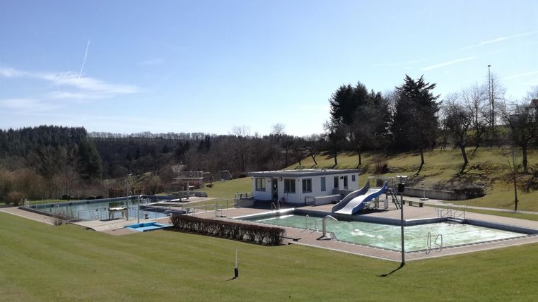 Ein hübsches Freibad mit mehreren Schwimmbecken und einer Rutsche. Im Hintergrund sind grüne Wiesen und Bäume sichtbar.