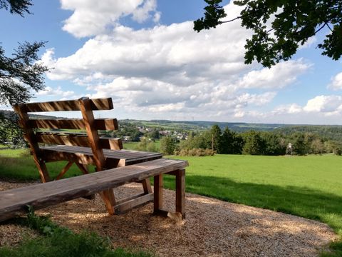 A large wooden bench sits on a hill overlooking a green landscape and a cloudy sky.