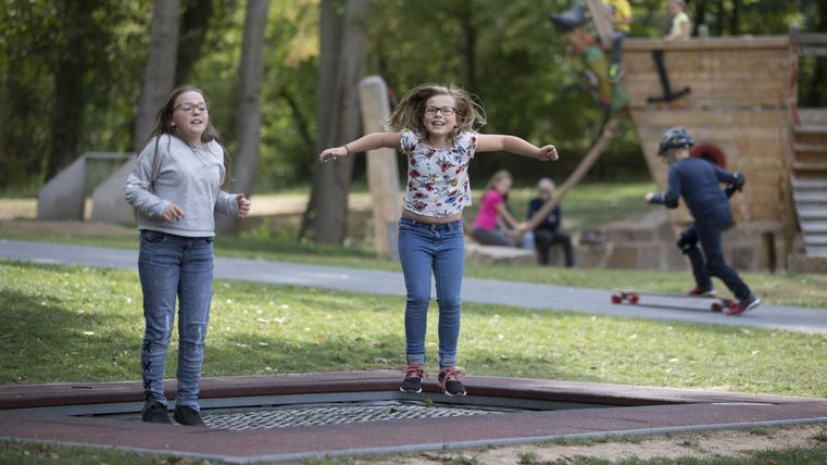Two cheerful girls are jumping on a trampoline in the park. In the background, more children are playing on a playground.