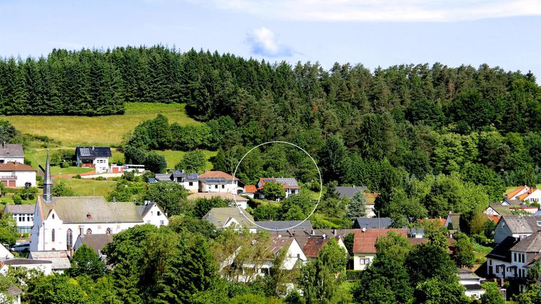 Eine ruhige ländliche Landschaft mit Wohnhäusern und viel Grün. Im Hintergrund sind Bäume und sanfte Hügel sichtbar.