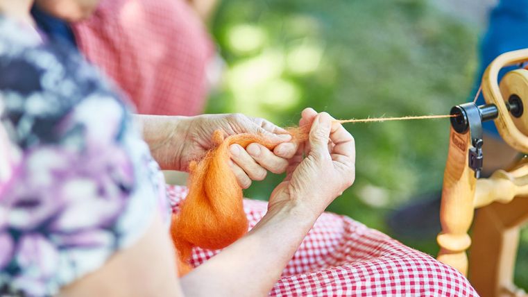 A person is spinning orange wool with a spinning wheel. In the background, more people can be seen.
